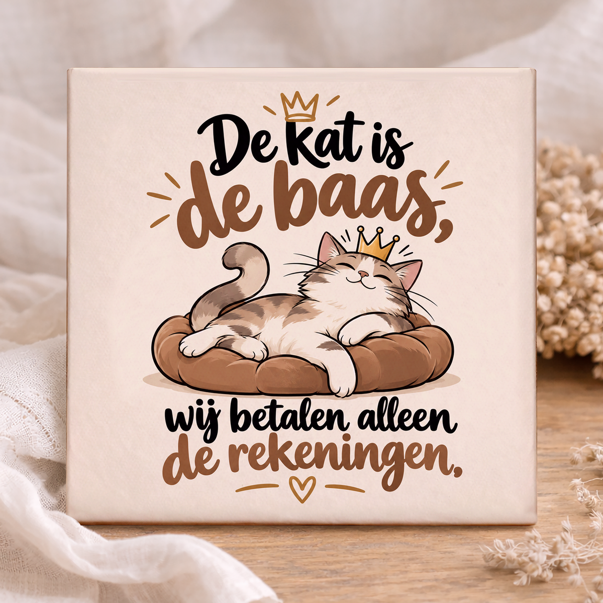 Keramisch katten tegeltje met tekst van 11x11 cm met tekst De kat is de baas wij betalen alleen de rekeningen. Grappige illustratie van een tevreden kat met gouden kroontje op een bruin kussen. Beige woondecoratie voor kattenliefhebbers.